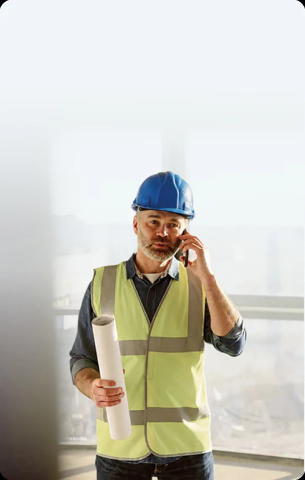 Bauleiter mit Schutzhelm und Warnweste auf der Baustelle, im Hintergrund eine Baustelle mit Fensterfront